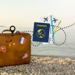A vintage brown suitcase decorated with travel stickers sits on a pebble beach beside the ocean, with a floating passport, plane tickets, and airplane graphic symbolizing travel and adventure.